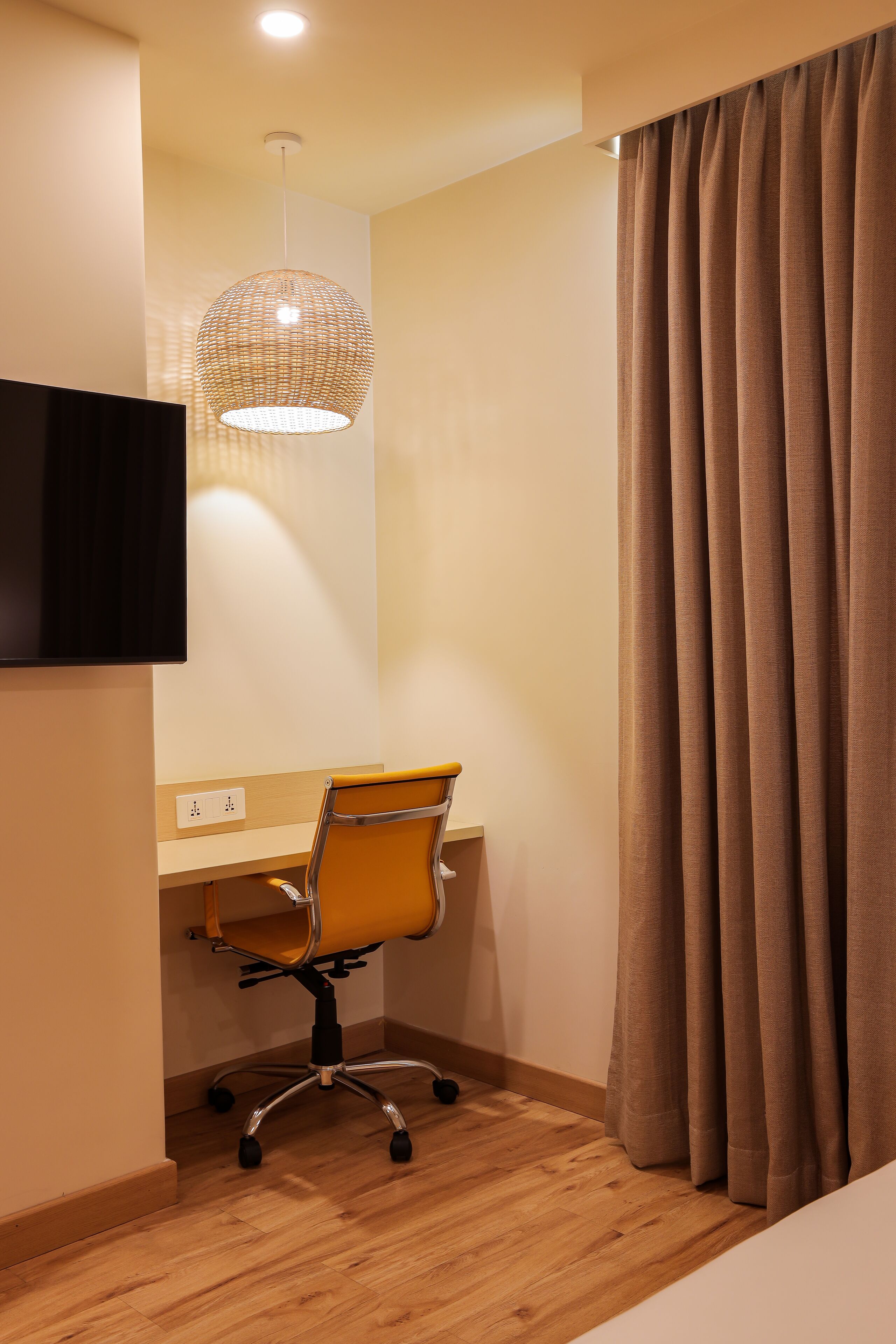 A modern hotel room with a wooden desk, a chair, a television mounted on the wall, and a large window with curtains.
