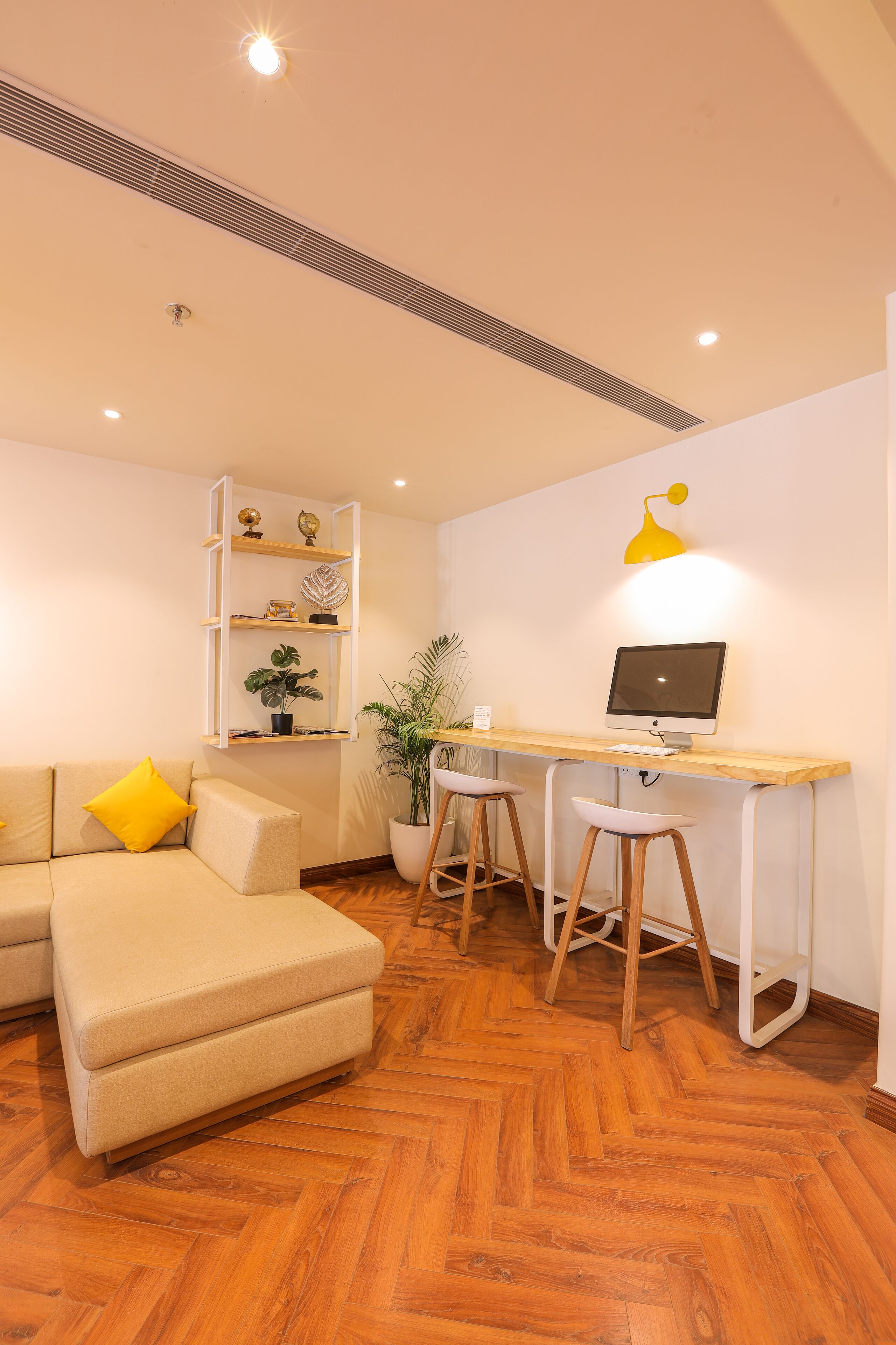 A modern living room with a wooden herringbone floor, a beige sofa with yellow cushions, a wooden desk with a computer, and a potted plant.