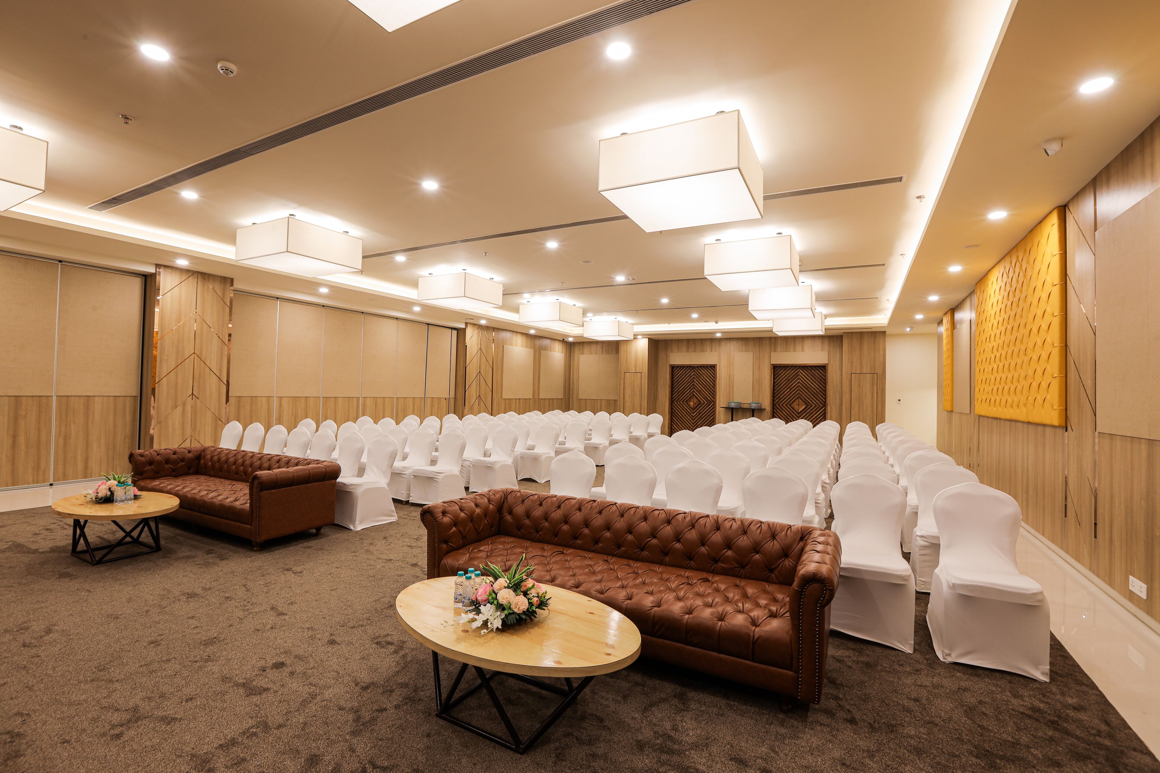 Un salón de conferencias con filas de sillas blancas, un sofá marrón y dos mesitas con centros de mesa florales.