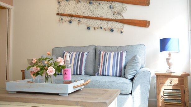 Een woonkamer met een bank, kussens, een houten bijzettafeltje, een lamp, een dienblad met bloemen en een decoratie aan de muur.