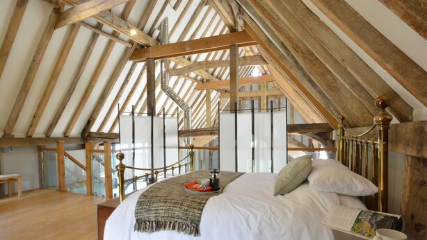 A high-ceilinged bedroom with a wooden beam structure, a bed with a white comforter, pillows, a bedside table with a book and a mug, and a view of the outside through a large window. - Manor House Barn, Peasenhall (Saxmundham)