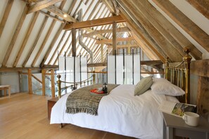 Une chambre avec un plafond haut et une structure en poutres apparentes, un lit recouvert d’une couette blanche, des oreillers, une table de chevet avec un livre et une tasse, et une vue sur l’extérieur par une grande fenêtre.