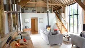 A spacious living area with a fireplace, wooden beams, a sofa, and a dining table with bread and vegetables. - Manor House Barn, Peasenhall (Saxmundham)