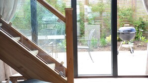 A glass door leading to an outdoor patio with a wooden staircase, a barbecue grill, and a garden view. - Manor House Barn, Peasenhall (Saxmundham)