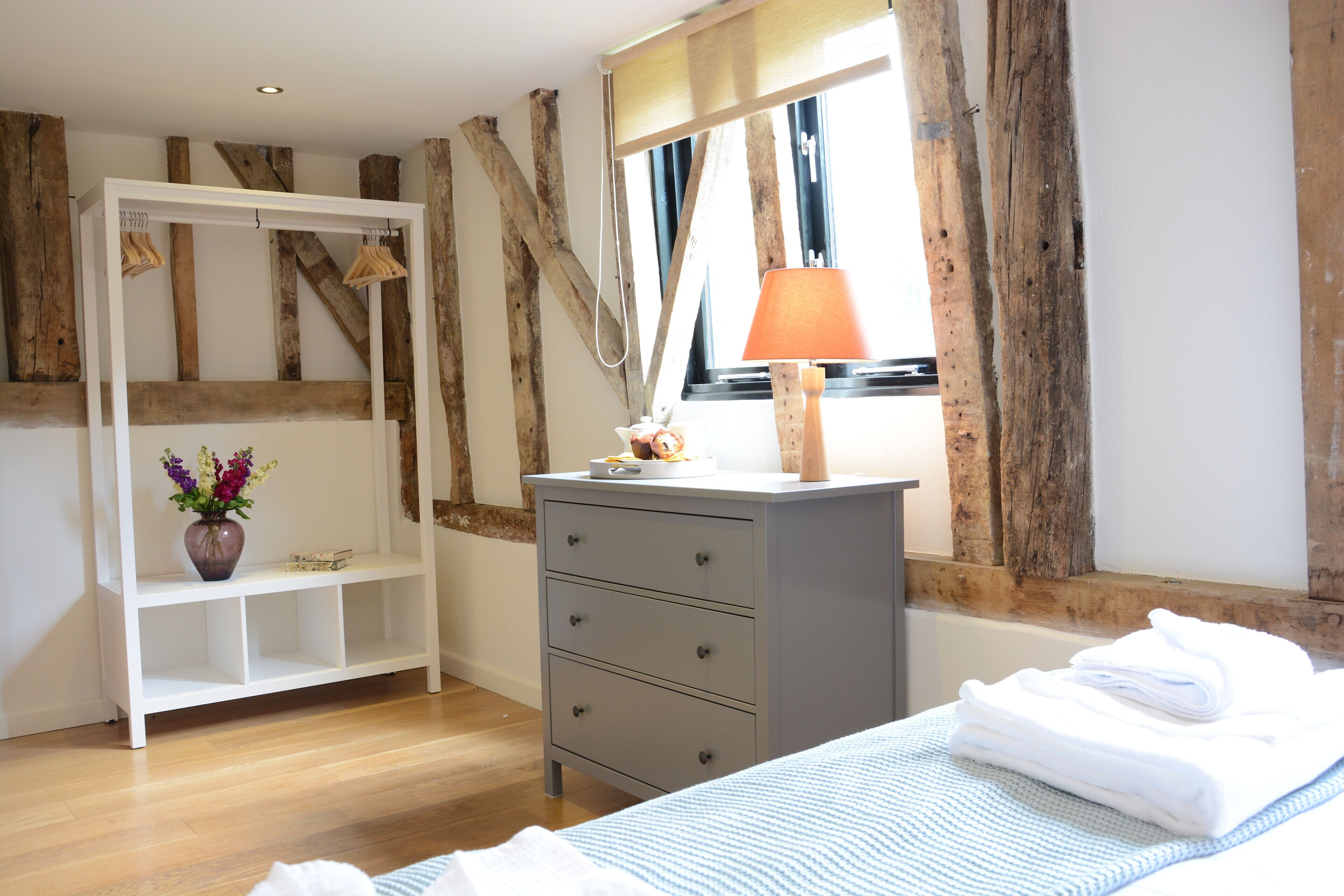 Un dormitorio con cama, cómoda, tocador, ventana y estructura de vigas de madera.
