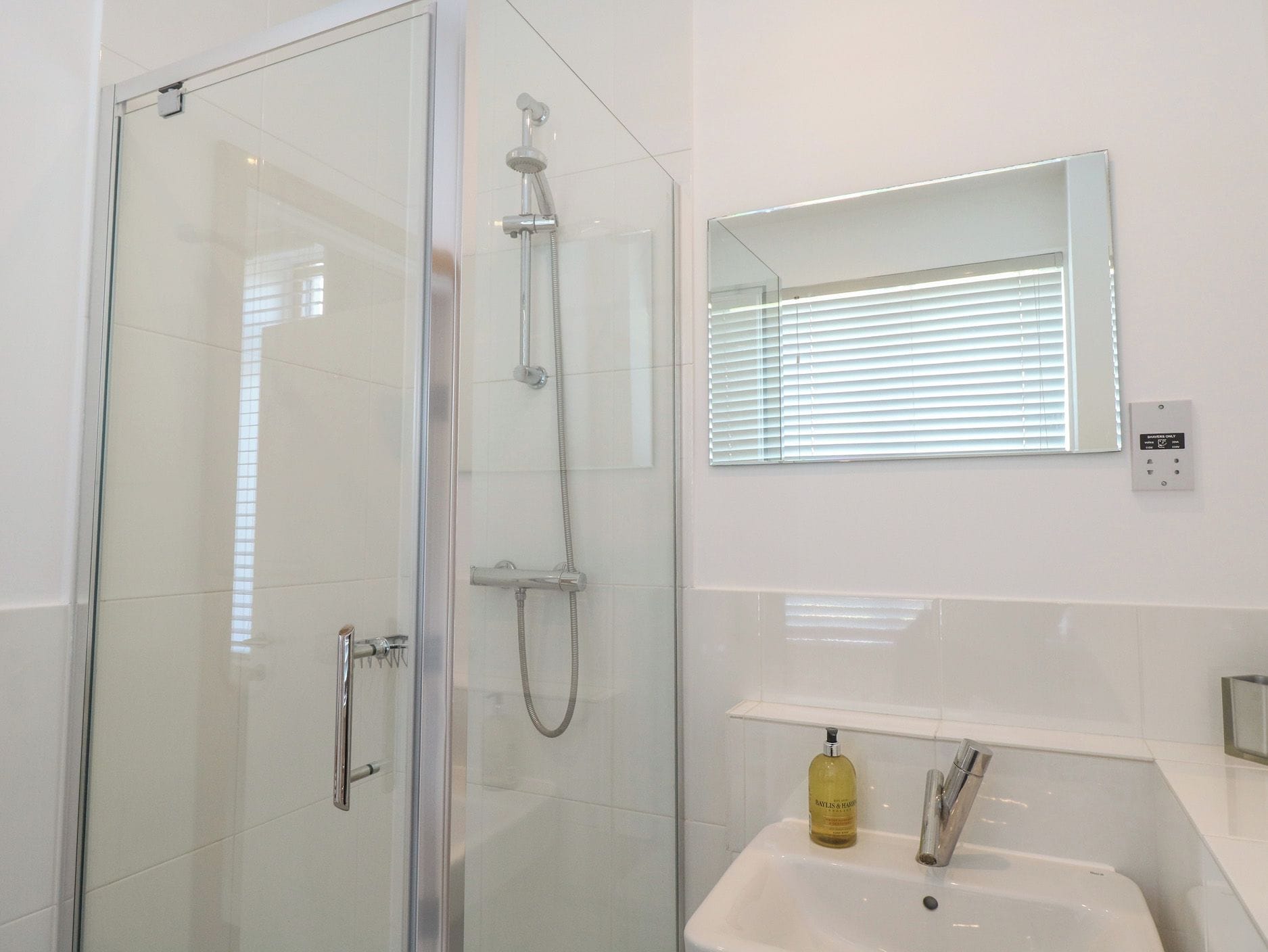 Un baño moderno con ducha envuelta en vidrio, un lavamanos blanco y un espejo sobre otro espejo.