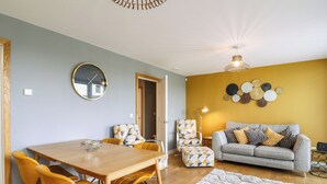 Un salon moderne avec un coin repas, comprenant une table à manger en bois, des chaises jaunes, un canapé gris agrémenté de coussins à motifs et un mur décoratif orné d’œuvres d’art murales de forme circulaire.