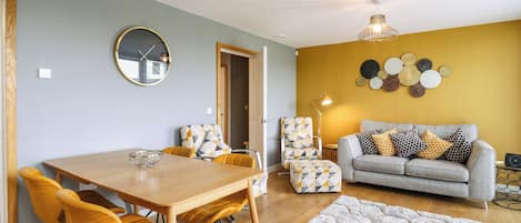 A modern living room with a dining area, featuring a wooden dining table, yellow chairs, a grey sofa with patterned cushions, and a decorative wall with circular wall art.
