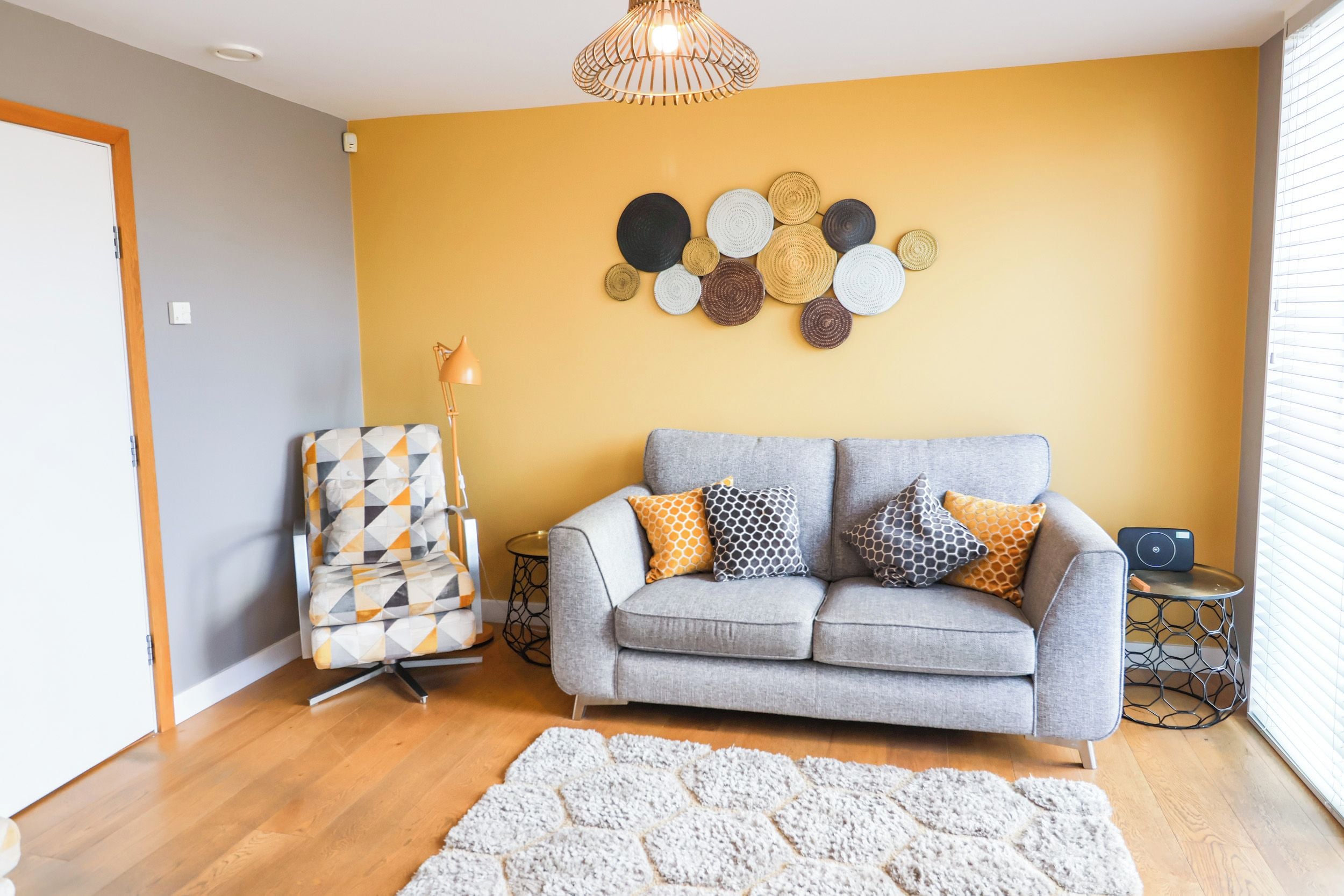 A living room with a grey sofa, patterned cushions, a geometric chair, a wall art piece, and a floor lamp.
