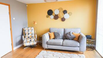 A living room with a grey sofa, patterned cushions, a geometric chair, a wall art piece, and a floor lamp.