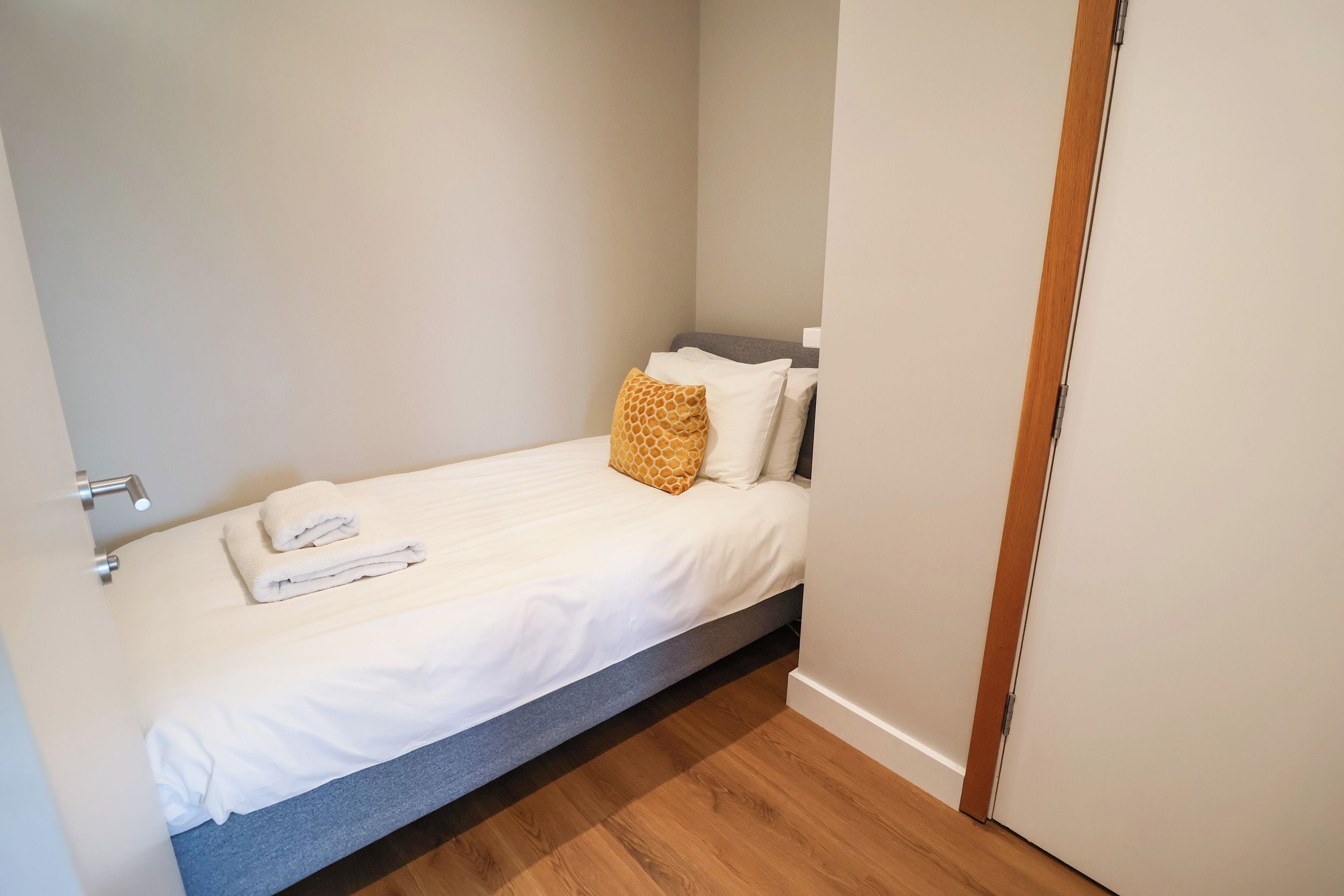 A small, single-bed room with a wooden floor, a grey headboard, and a white bedspread.