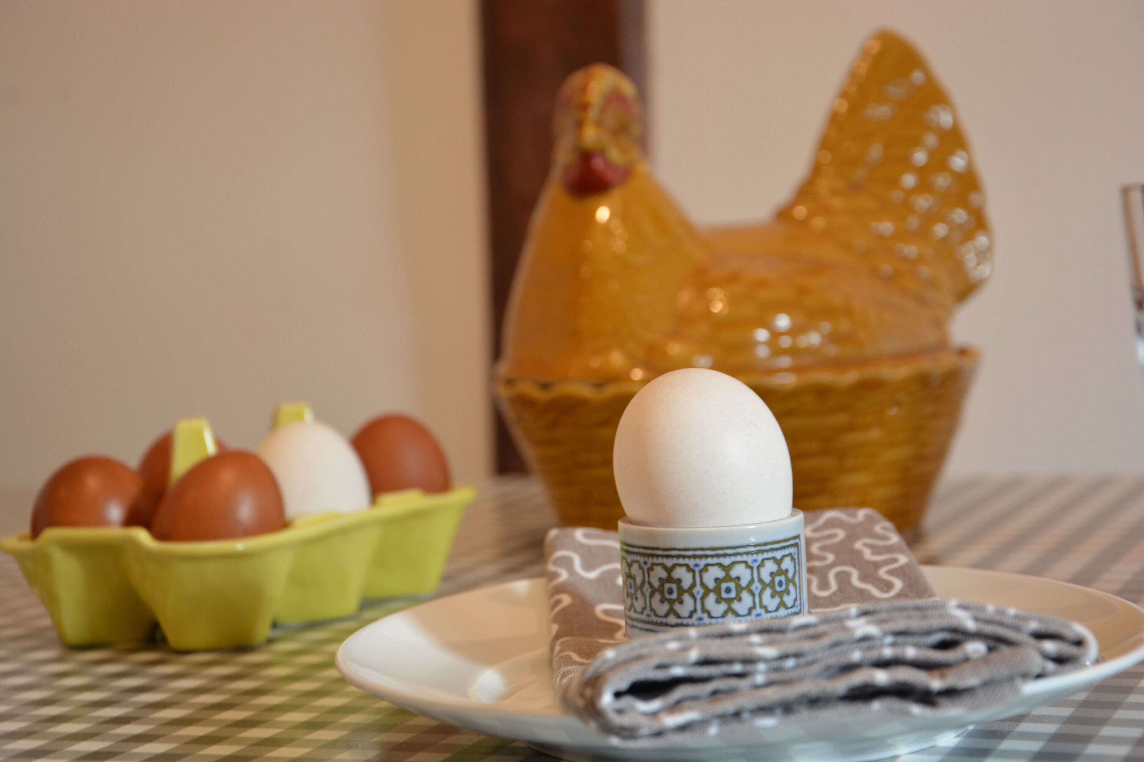 A table with a checkered tablecloth, a plate with a rolled-up item, a ceramic chicken, and a carton of eggs.