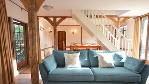 A living room with a blue sofa, wooden beams, a staircase, and a dining area. - Wheelwright's Barn (Halesworth)