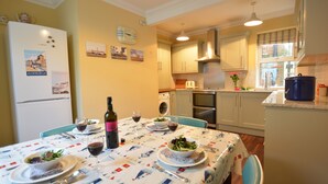 A dining area with a table set for two, a refrigerator, a washing machine, and a kitchen in the background. - Gosfield Cottage, Aldeburgh (Aldeburgh)