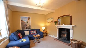 A cozy living room with a blue sofa, a fireplace, a wooden table, and a wicker basket. - Gosfield Cottage, Aldeburgh (Aldeburgh)