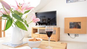 Ett mysigt vardagsrum med en tv, en skål med snacks, ett glas rött vin, en bok och en vas med blommor.