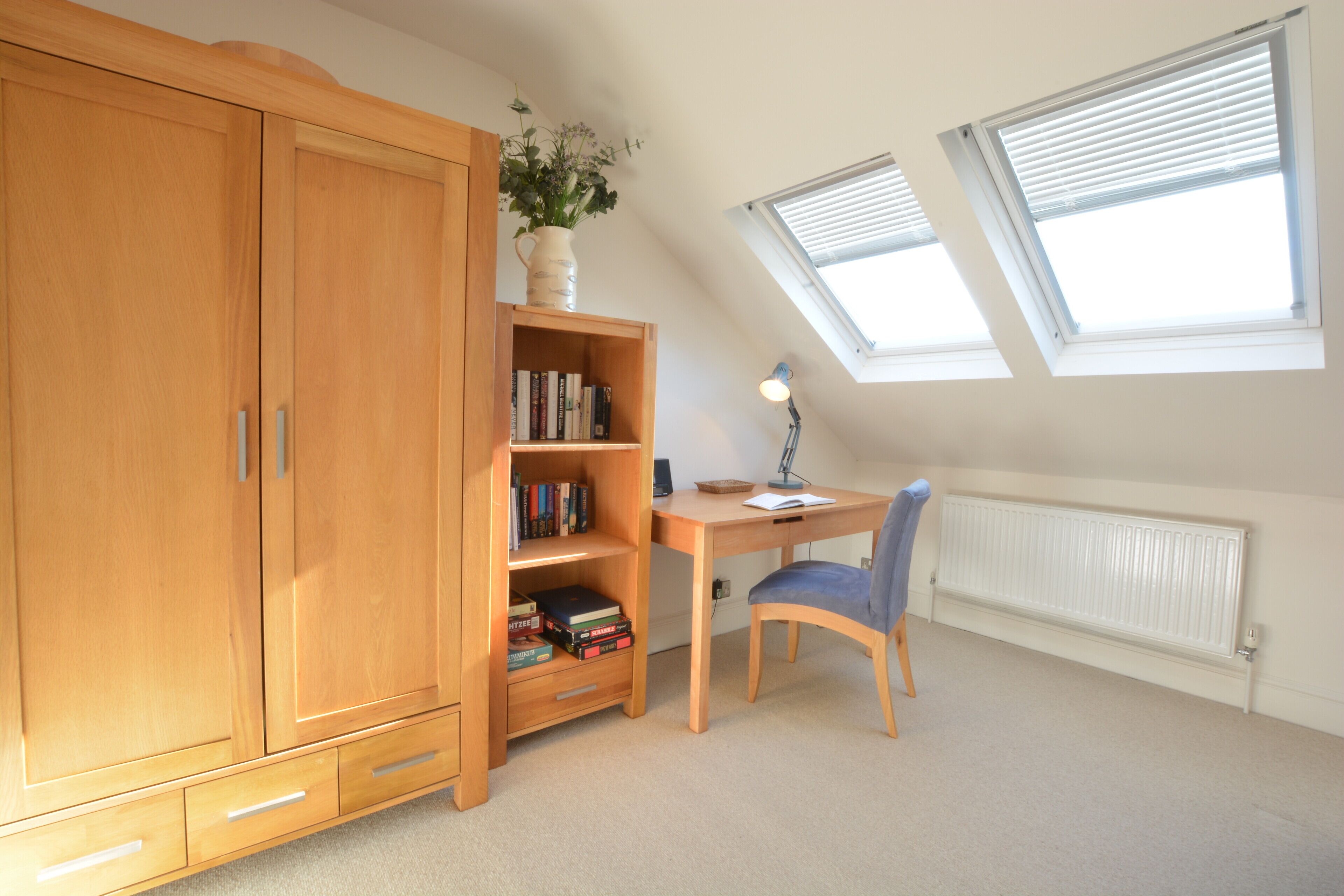 Een kamer met een houten garderobe, een boekenkast vol boeken, een bureau met een stoel, en een radiator. Er zijn twee dakramen in het plafond.