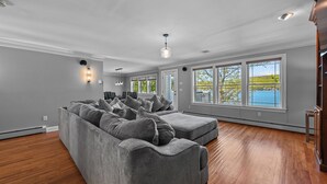 A spacious living room with a large sectional sofa, multiple windows offering a view of a lake, and hardwood flooring.