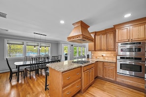 A modern kitchen with wooden cabinets, a granite countertop, and stainless steel appliances. - Lakefront Paradise | Dock • Hot Tub • Sleeps 10 (West Milford)