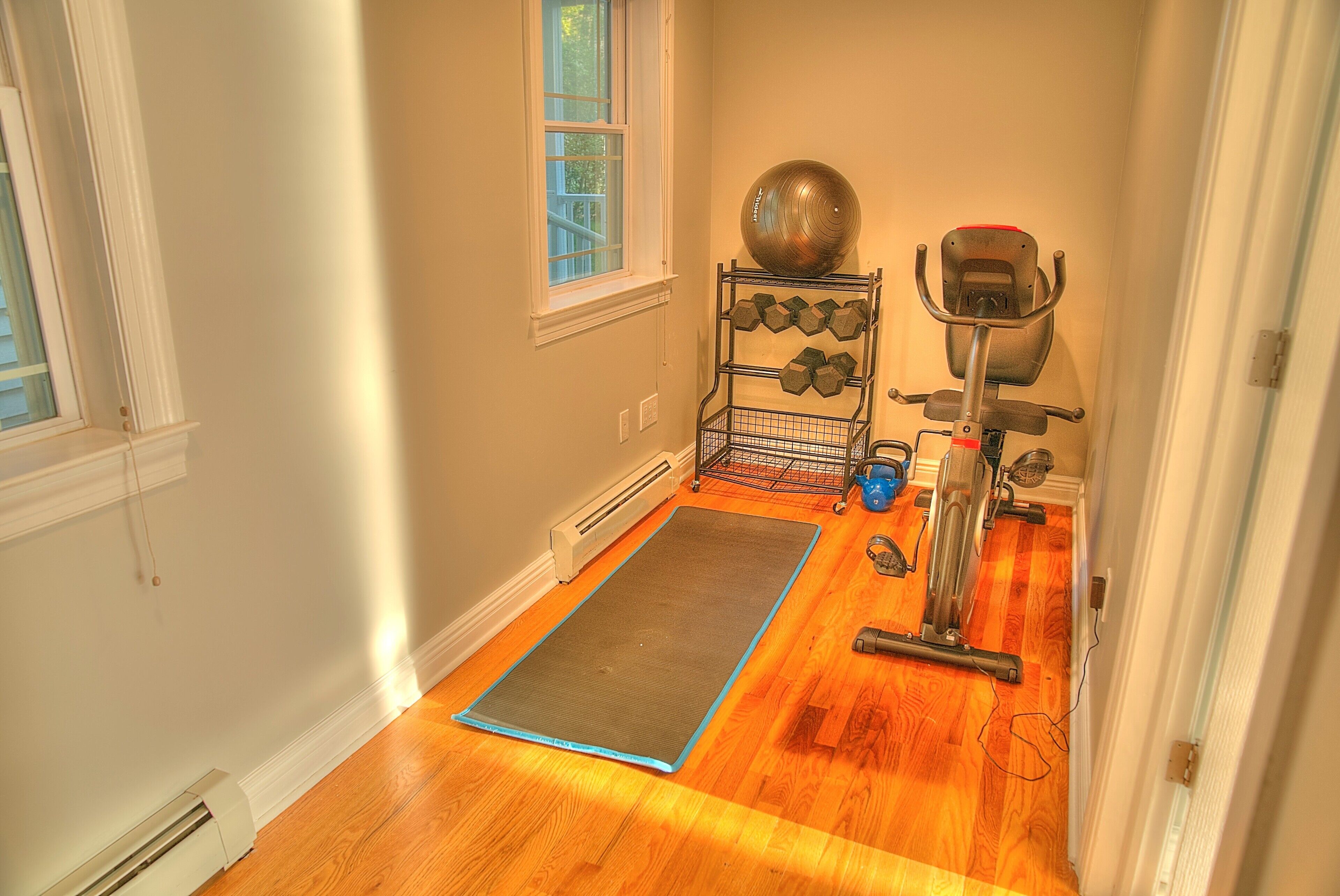 A room with a wooden floor, a yoga mat, a stationary bike, a set of dumbbells, and a large exercise ball.