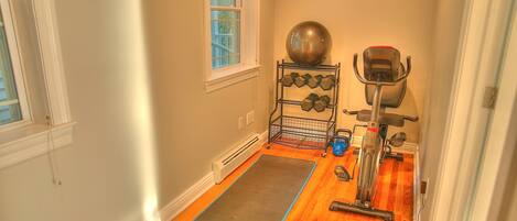 A room with a wooden floor, a yoga mat, a stationary bike, a set of dumbbells, and a large exercise ball.