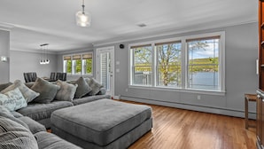 A spacious living room with a large sectional sofa, a grey ottoman, and a view of a lake through large windows.