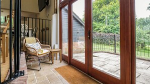 A cozy room with a wicker chair, a small wooden table, and a large sliding glass door leading to a patio with a brick wall and a metal fence. - Quill Farm Barn (Woodbridge)