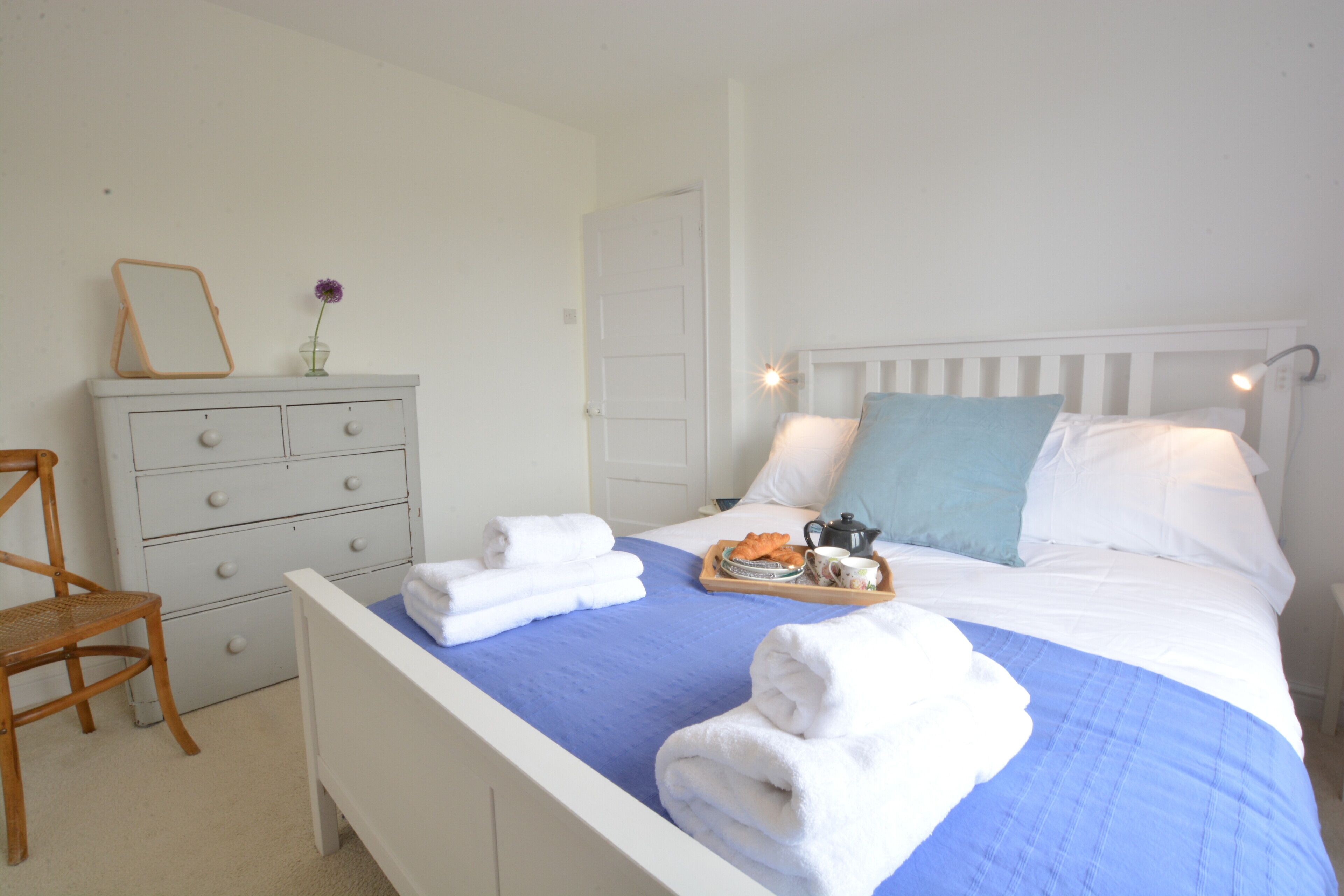 A neatly made bed with a blue bedspread, white pillows, and a tray with towels and a teapot.