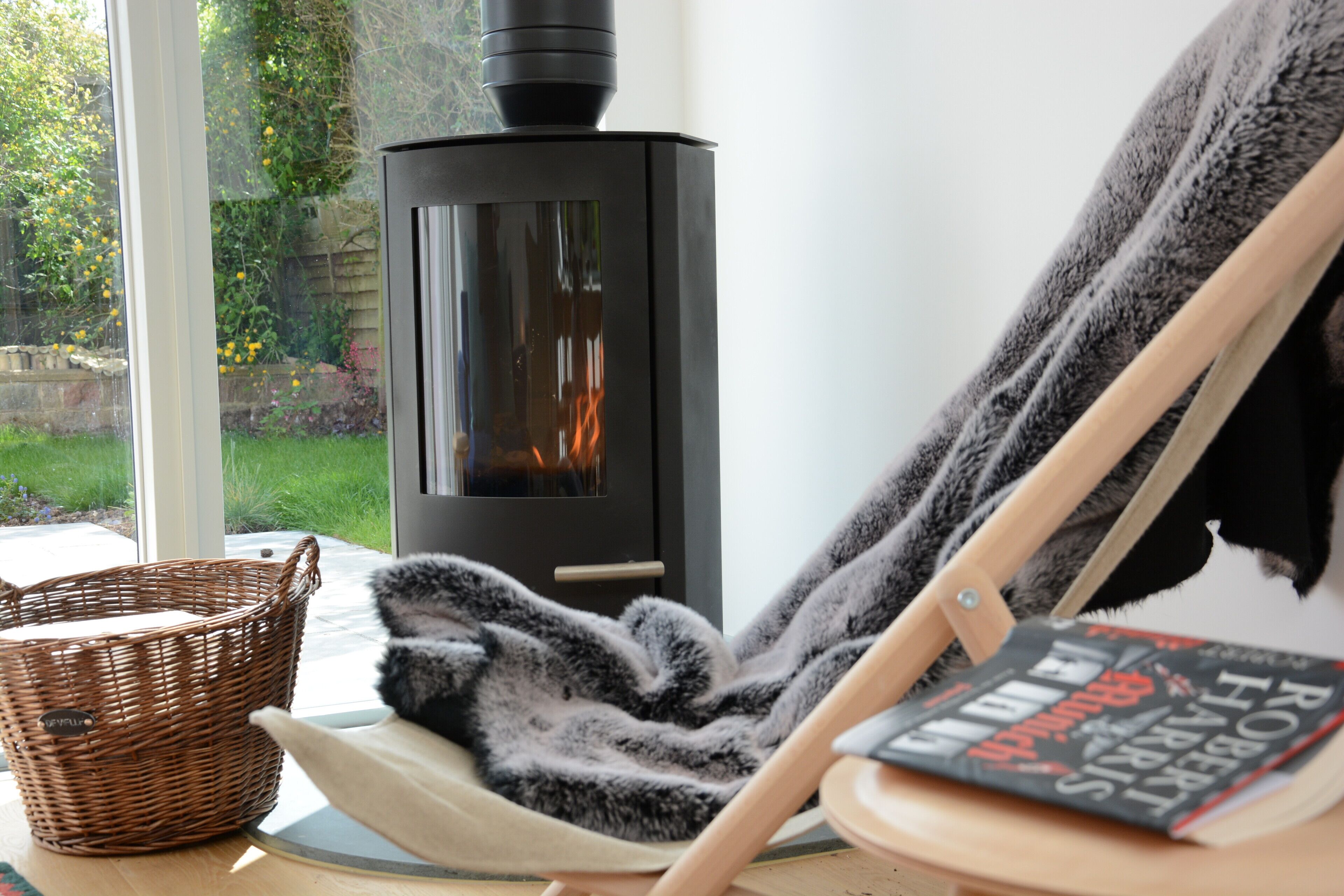 A cozy room with a wood-burning stove, a wicker basket, a chair with a blanket, and a book on a wooden stand.