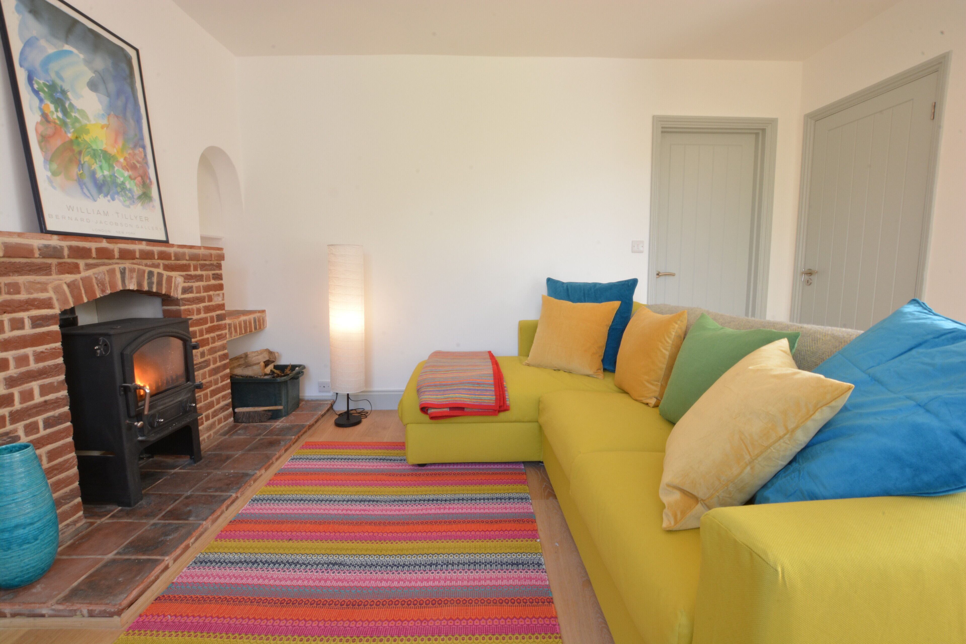 A living room with a brick fireplace, a yellow sofa with colorful cushions, a striped rug, and a framed picture on the wall.