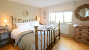 A bedroom with a wooden bed frame, a bedside table with a lamp, a dresser, and a window with a view. - The Nookery at Snape Hall (Saxmundham)