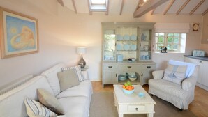 A living room with a white sofa, a striped armchair, and a coffee table with fruit. There is a cabinet with plates and bowls, and a painting on the wall. - The Nookery at Snape Hall (Saxmundham)