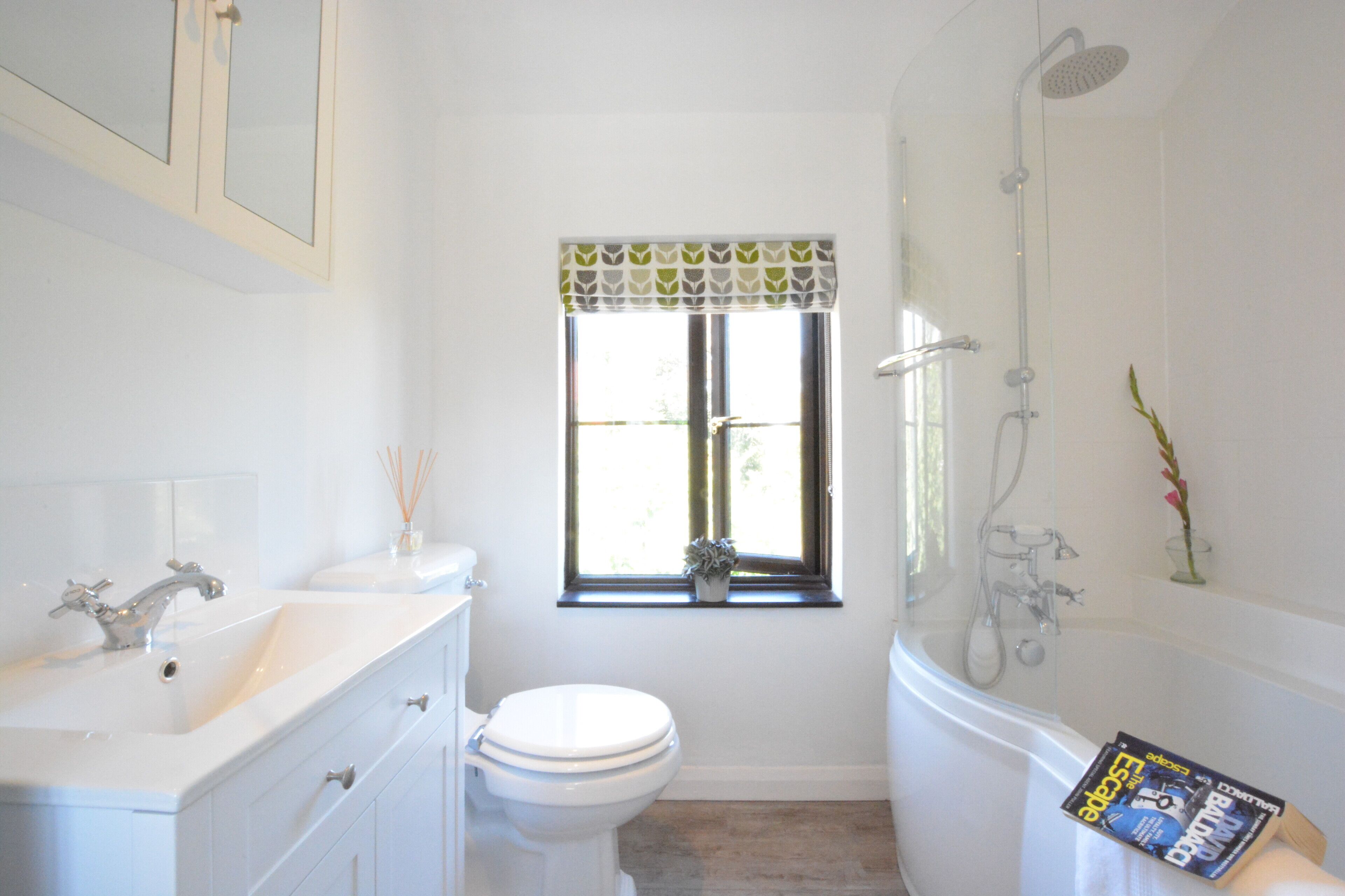 Un baño con lavamanos, inodoro y bañera blancos. Se ve una ventana con un adornos estampados, y una ducha a la derecha.