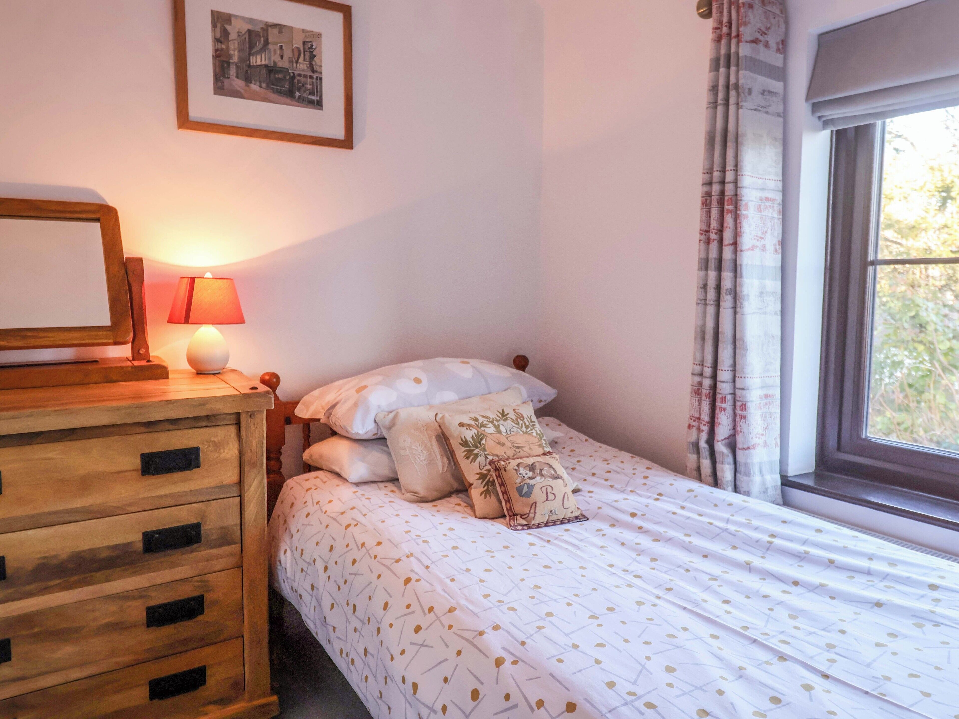 Un dormitorio con una cama, un aparador de madera con cajones, un cuadro enmarcado en la pared y una ventana con cortinas.