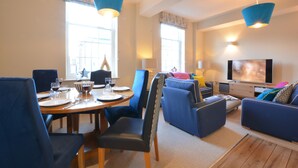 A modern living room with a dining area, featuring blue upholstered furniture, a wooden dining table, and a flat-screen TV. - Skimming Stones, Aldeburgh (Aldeburgh)