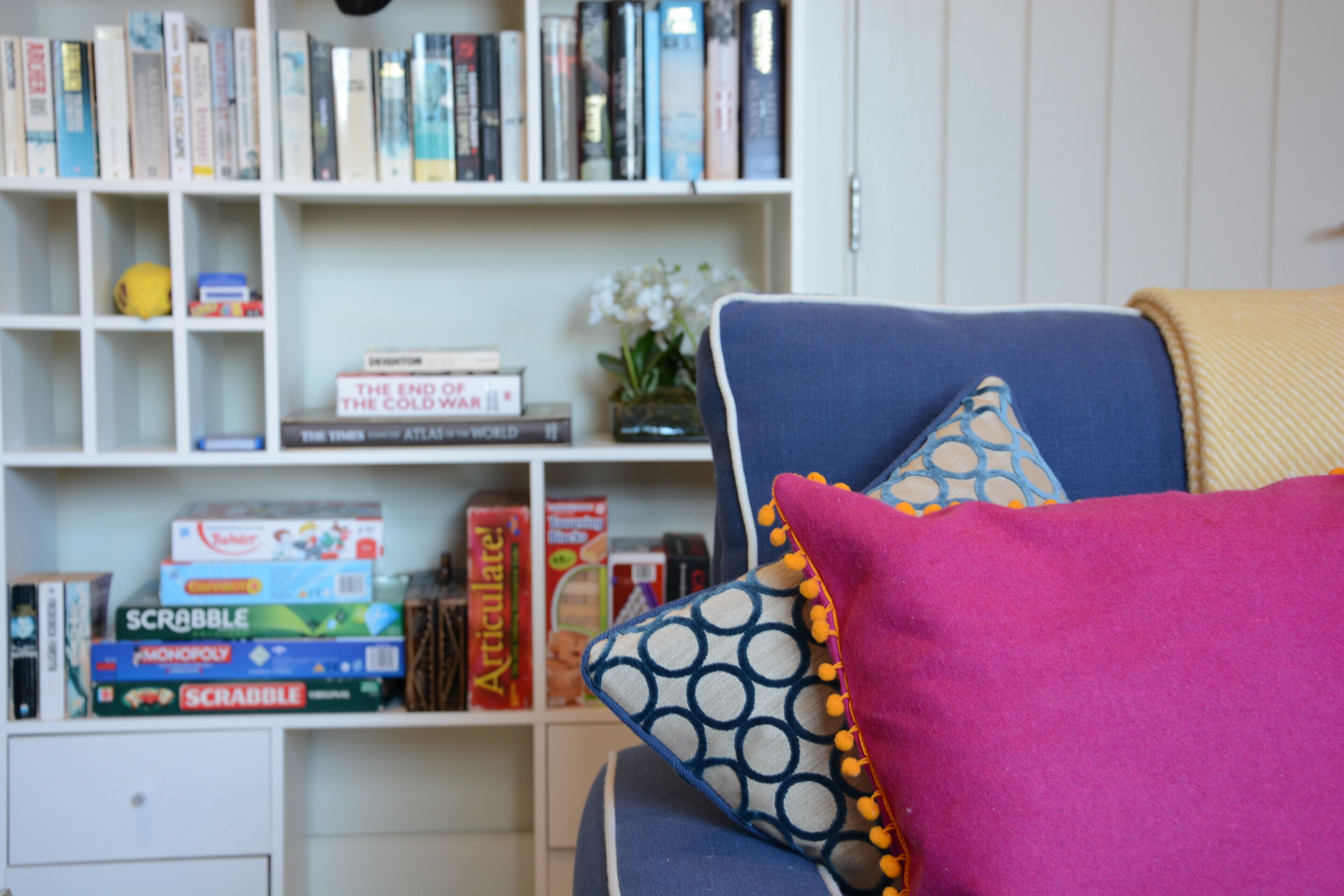 A bookshelf filled with books and board games, a blue sofa with colorful pillows, and a small vase with white flowers.