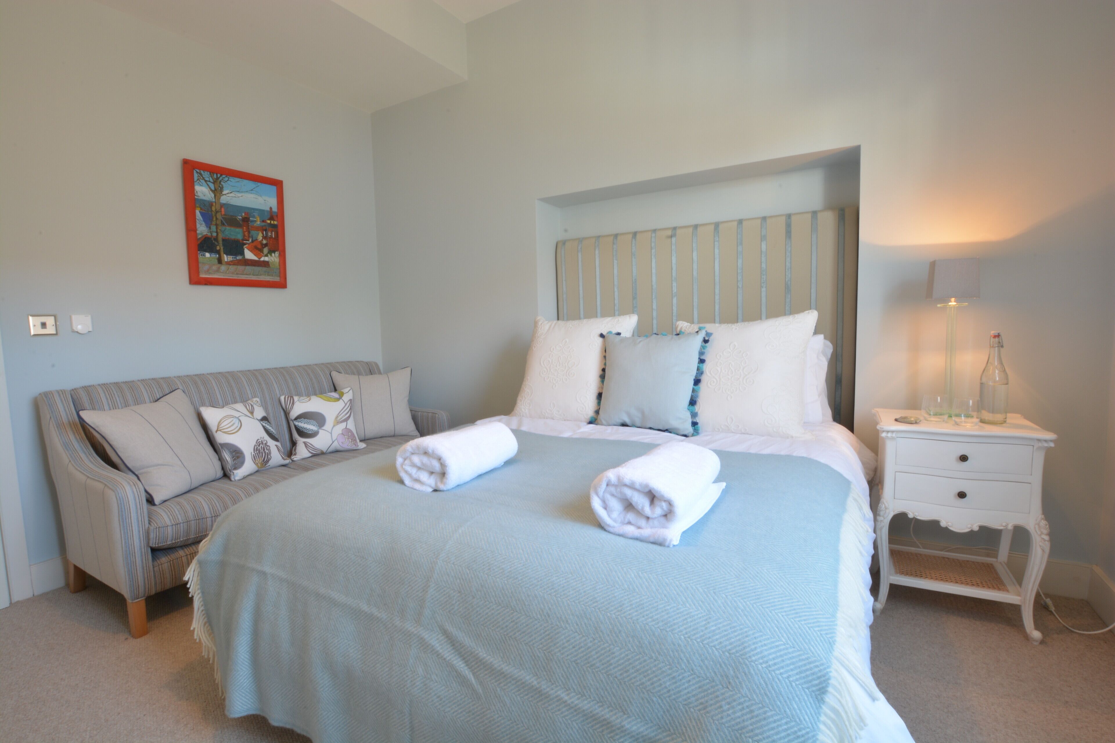 A neatly made bed with two rolled towels, a bedside table with a lamp, and a couch with pillows.