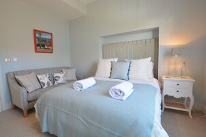 Cama bem arrumada com duas toalhas enroladas, mesa de cabeceira com abajur e sofá com almofadas.