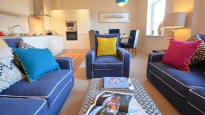 A modern living room with a sofa, armchair, and ottoman, a dining area with chairs, and a kitchen in the background. - Skimming Stones, Aldeburgh (Aldeburgh)