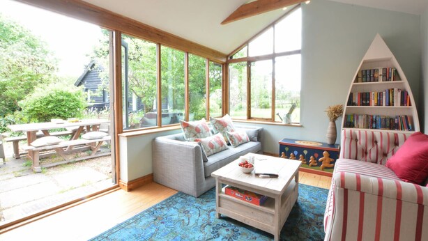 A cozy living room with a large window, a sofa, a striped armchair, a bookshelf, and a wooden coffee table.
