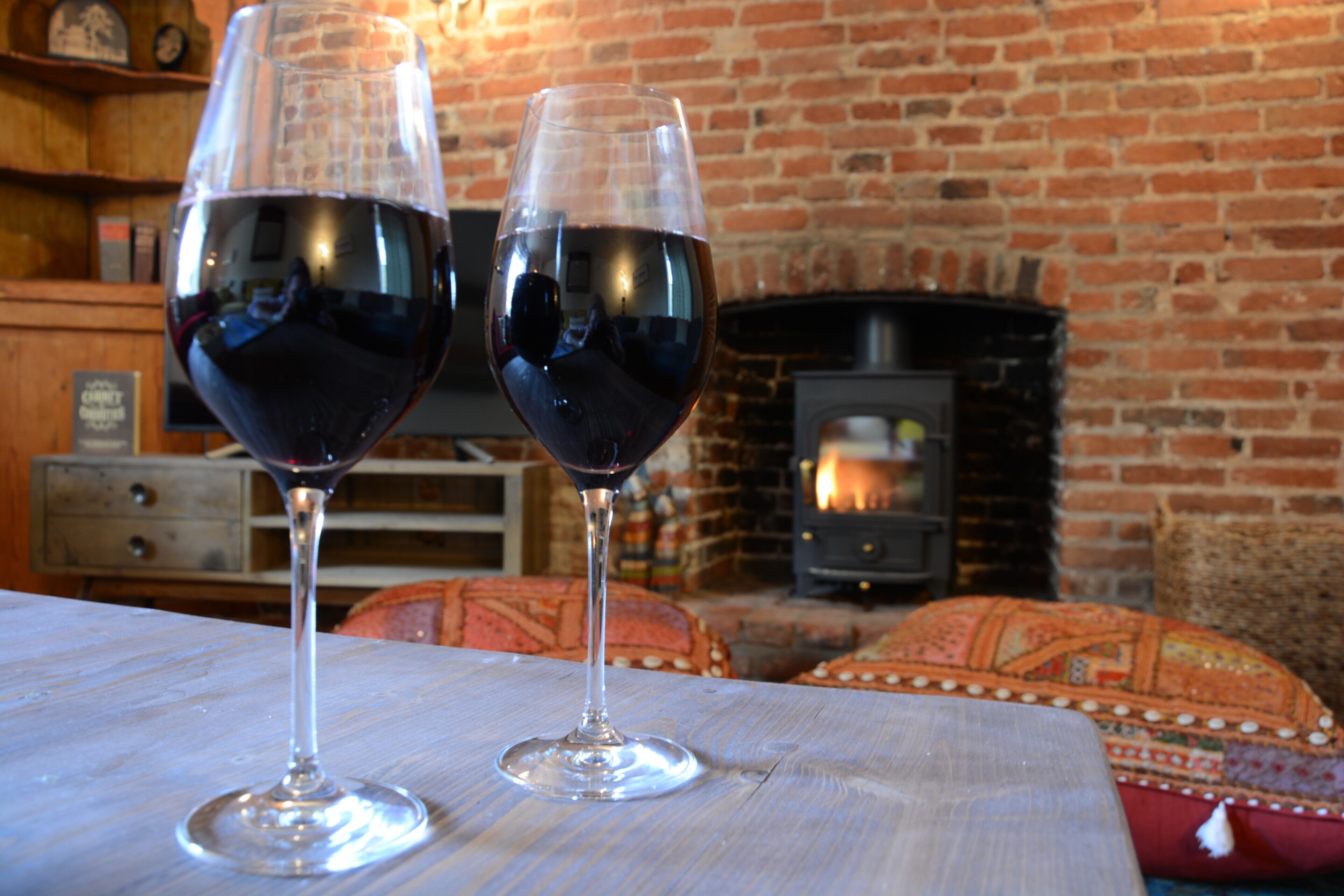 Deux verres à vin sont posés sur une table en bois dans une pièce dotée d’une cheminée en briques.