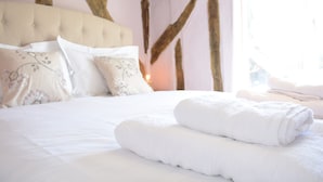 A neatly made bed with white linens and pillows, a wooden headboard, and a window with daylight coming through.