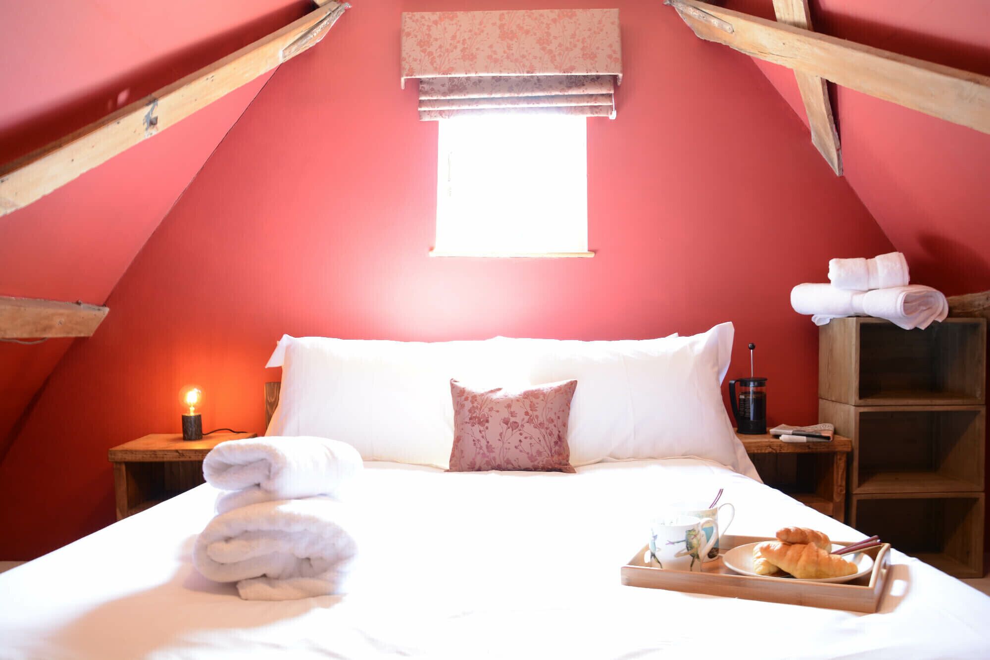 Un dormitorio acogedor con techo inclinado, una cama con sábanas blancas, una mesita de noche con una lámpara y una bandeja con cruasanes y una taza.