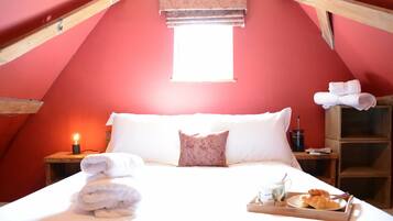 Un dormitorio acogedor con techo inclinado, una cama con sábanas blancas, una mesita de noche con una lámpara y una bandeja con cruasanes y una taza.