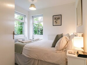A bedroom with a bed, a bedside table with a lamp, a framed picture on the wall, and windows with a view of greenery. - The Bridge House, Southwold (Reydon)