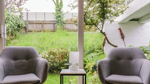 Two grey armchairs facing a window with a view of a garden and a white brick wall. - The Bridge House, Southwold (Reydon)