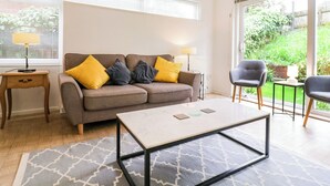 A living room with a sofa, two chairs, a coffee table, and a view of a garden through large windows. - The Bridge House, Southwold (Reydon)