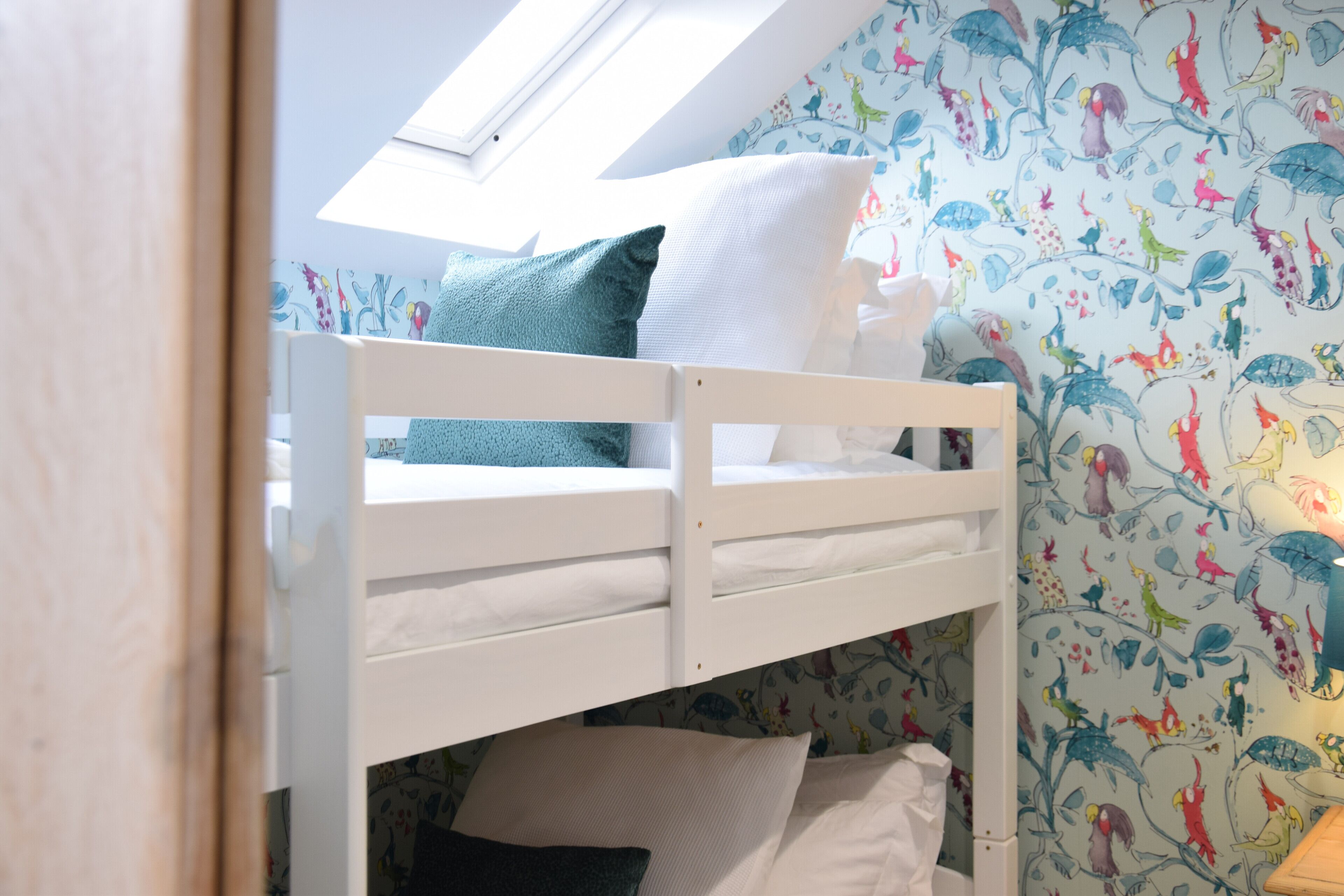 A bunk bed with a skylight above, white bedding, and a wall with colorful bird and plant wallpaper.