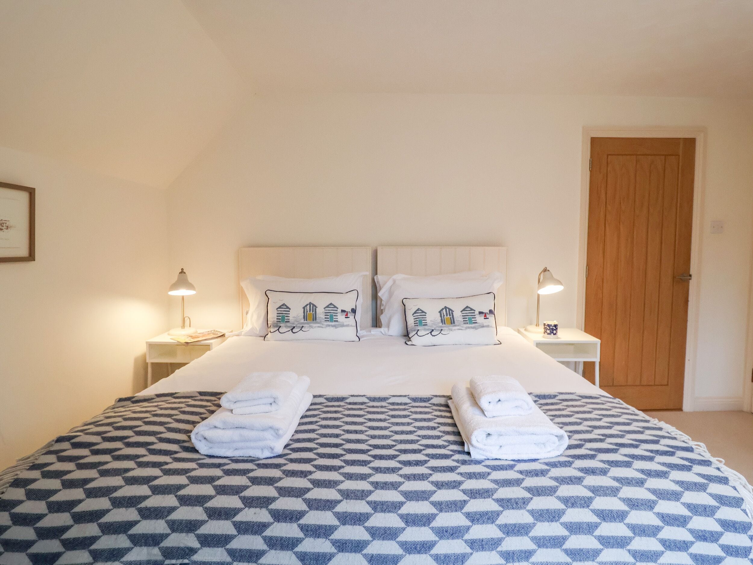 Una cama bien hecha con colcha a cuadros, dos almohadas con diseño de paisaje costero y dos toallas dobladas. Se observan dos lámparas de noche, un cuadro enmarcado en la pared y una puerta de madera al fondo.