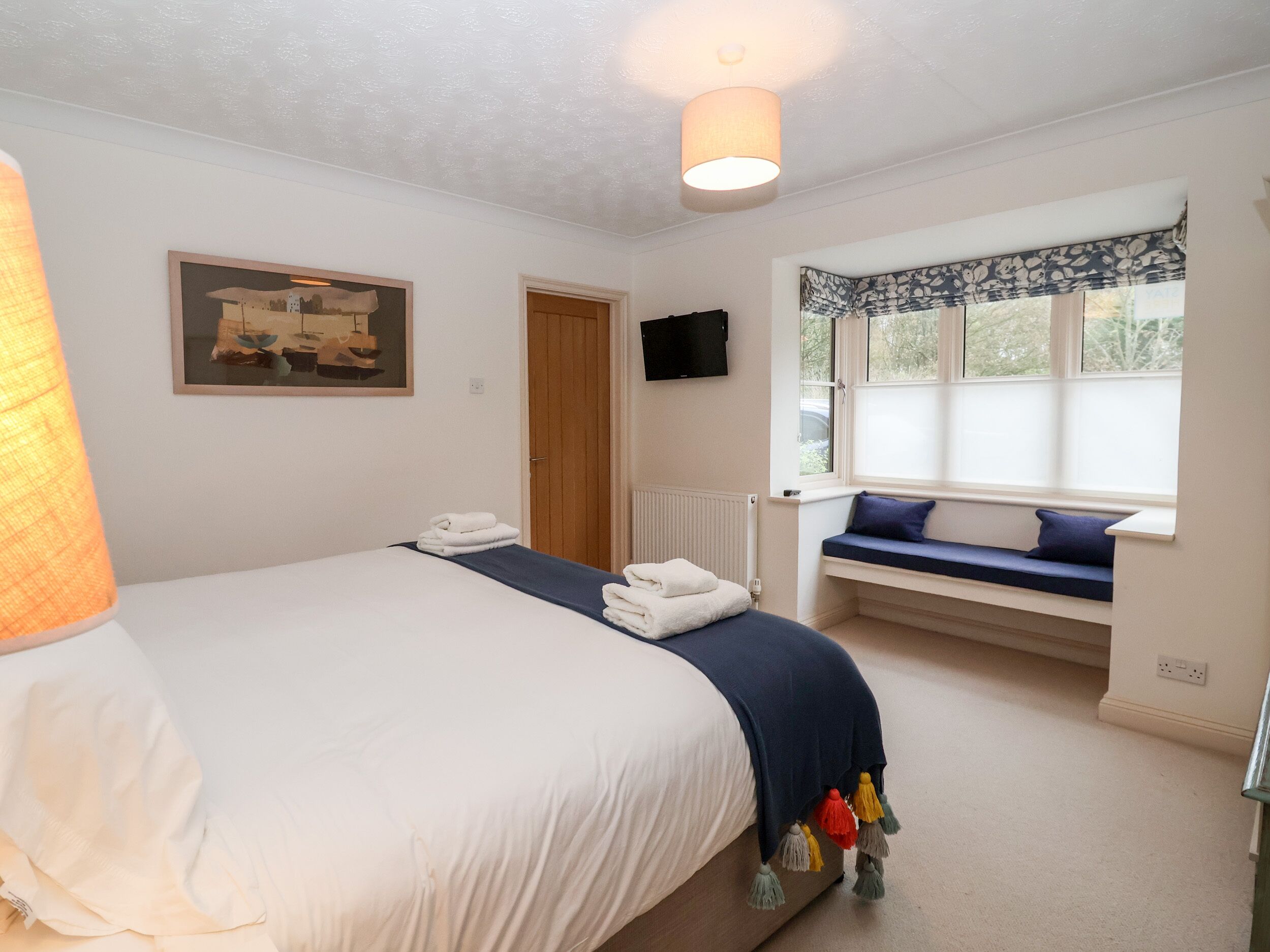A bedroom with a bed, a window seat, a painting, and a television on the wall.